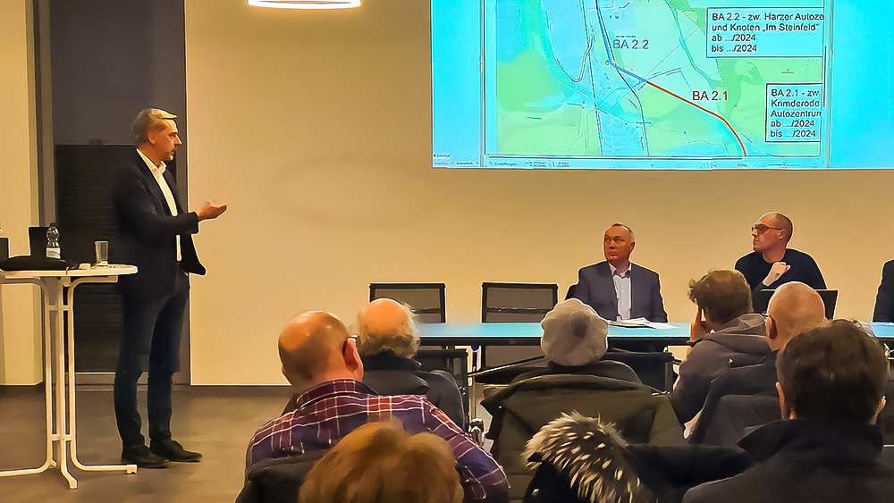 Ein Mann erklärt in einer Präsentation mit Beamer eine Karte, zwei weitere Männer sitzen im Plenum, Zuhörer im Vordergrund.
