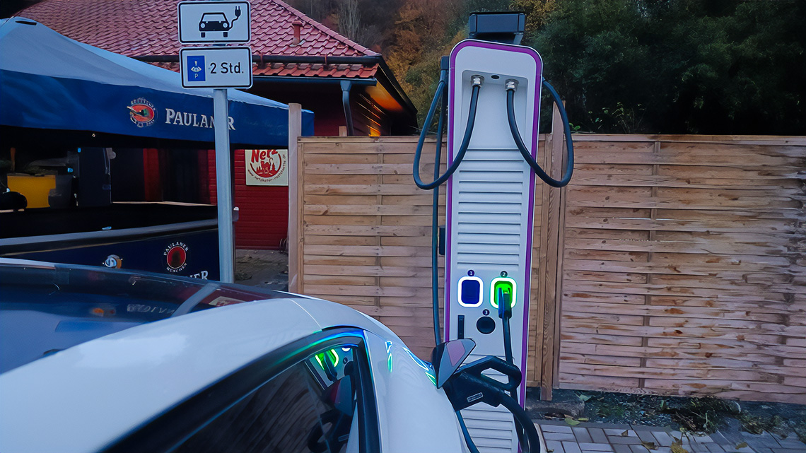 Eine Ladestation für elektrische Fahrzeuge steht vor einem Holzzaun an der Raststätte Netzkater im Südharz. Ein weißer PKW ist angeschlossen und lädt.