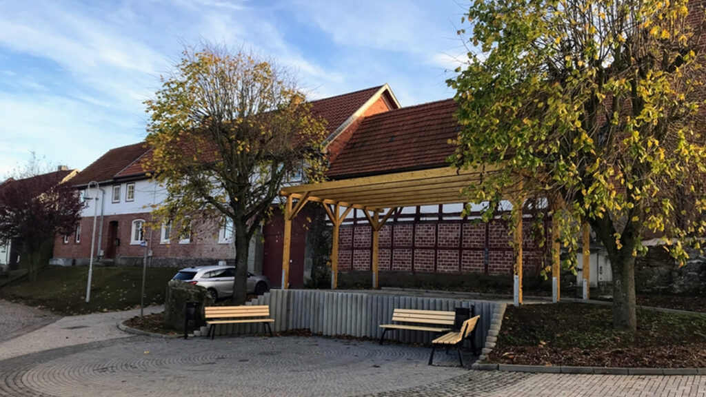 Bauernhäuser hinter einem Dorfplatz, darauf ein Überdachung und drei Sitzbänke.