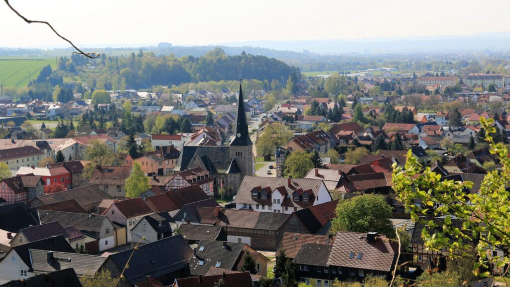 Eine Stadt mit vielen Häusern und einer Kirche mit einem hohen Kirchturm in der Mitte. Die Stadt ist von Bäumen und Hügeln im Hintergrund umgeben.