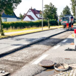 Bild von Straßenbauarbeiten an einer Ortsstraße mit Baumaschine, im Hintergrund eine grüne Hecke.