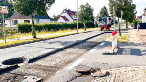 Bild von Straßenbauarbeiten an einer Ortsstraße mit Baumaschine, im Hintergrund eine grüne Hecke.
