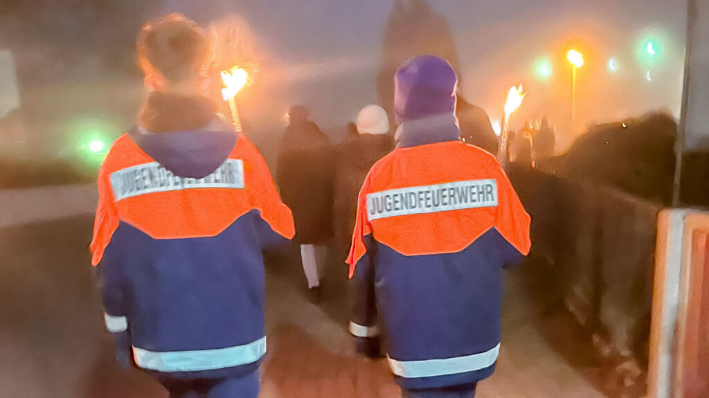 Zwei Mitglieder der Jugendfeuerwehr, von hinten fotografiert, mit Fackeln in der Hand, gehen durch den nebligen Ort.