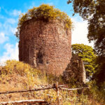 Ein alter, verfallener Turm steht im Freien. Er ist teilweise von Grünflächen und Bäumen umgeben, die in warmen Herbstfarben leuchten. Ein Holzzaun führt zum Turm, und der Hintergrund zeigt einen klaren blauen Himmel mit einigen Wolken. Die Szene strahlt eine ruhige, historische Atmosphäre aus.