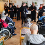Eine Gruppe älterer Menschen sitzt in Rollstühlen und Stühlen in einem hellen Raum, während eine Chorkapelle vor ihnen steht und singt. Die Sänger halten Notenbücher und sind engagiert bei der Performance. Der Raum ist freundlich eingerichtet mit Bildern an den Wänden und einer angenehmen Atmosphäre.