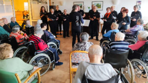 Eine Gruppe älterer Menschen sitzt in Rollstühlen und Stühlen in einem hellen Raum, während eine Chorkapelle vor ihnen steht und singt. Die Sänger halten Notenbücher und sind engagiert bei der Performance. Der Raum ist freundlich eingerichtet mit Bildern an den Wänden und einer angenehmen Atmosphäre.
