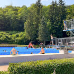 Menschen im Freibad vor bewaldeter Kulisse – Besucherinnen und Besucher schwimmen, sitzen am Beckenrand oder stehen am Sprungturm. Sommerliche Szene im Waldbad Ilfeld der Gemeinde Harztor. Gesichter wurden unkenntlich gemacht.