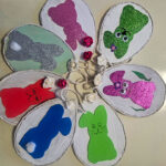 Bastelarbeit in Blumenform: Acht ovale Anhänger aus Pappe sind kreisförmig angeordnet und jeweils mit bunten Osterhasen aus Glitzerpapier verziert. In der Mitte verbinden kleine künstliche Blüten die Anhänger. Die Hasen sind in verschiedenen Farben gestaltet – darunter Grün, Pink, Silber, Blau und Rot. Die Dekoration liegt auf einem hellen Untergrund.