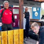 **ALT-Text:** Vier Männer und ein Kind arbeiten bei Sonnenschein gemeinsam an der Instandsetzung eines Holzgeländers vor dem Vereinsheim des SV Ilfeld. Im Hintergrund ist das Vereinslogo zu erkennen, die Stimmung wirkt freundlich und engagiert.