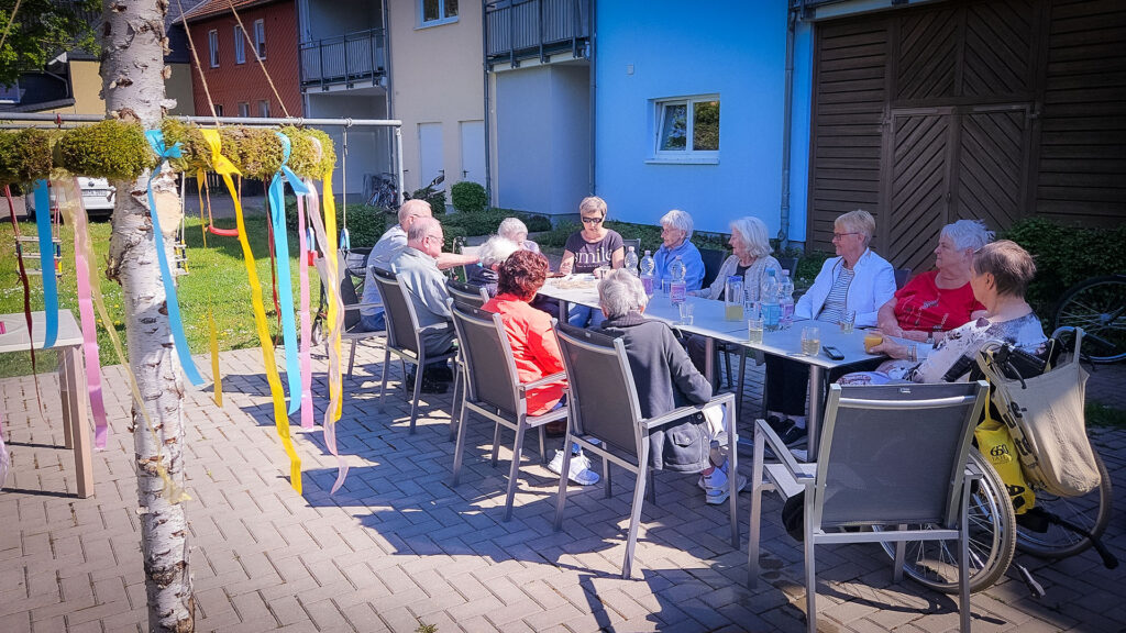 Eine Gruppe älterer Menschen sitzt an einem langen Tisch im Freien und genießt das Beisammensein bei Getränken.