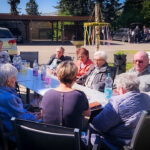 Eine Gruppe älterer Menschen sitzt an einem langen Tisch im Freien und genießt das Beisammensein bei Getränken.