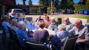 Eine Gruppe älterer Menschen sitzt an einem langen Tisch im Freien und genießt das Beisammensein bei Getränken.