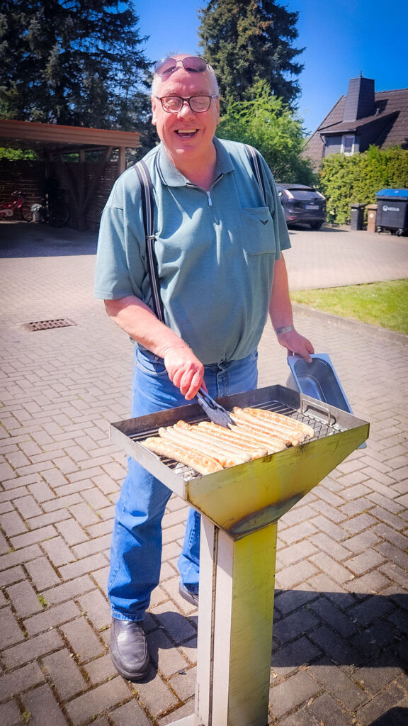 Ein lächelnder Mann grillt Bratwürste an einem Grill auf einem gepflasterten Hof.