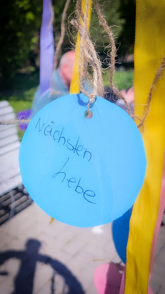 Nahaufnahme eines blauen Wunschkärtchens mit der handgeschriebenen Botschaft „Nächstenliebe“, befestigt an bunten Bändern.