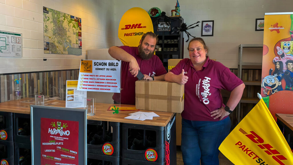 Zwei Mitarbeitende des Happylino Familien-Erlebnis-Centers präsentieren im Innenbereich einen neuen DHL-Paketshop mit Infotafel, Kartons und freundlichem Daumen-hoch.