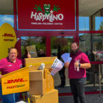 Zwei Mitarbeitende des Happylino Familien-Erlebnis-Centers stehen lächelnd mit DHL-Paketshop-Materialien und Paketen vor dem Eingang des Centers.