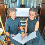 Das Duo Vimaris bei der Vorbereitung auf das Konzert an der Ladegast-Orgel in der St.-Georg-Kirche Neustadt/Harz: Dr. Wieland Meinhold zeigt auf eine Notenstelle, während Mirjam Meinhold mit einem Lächeln zuhört. Im Hintergrund die historische Orgel und der Blick ins Kirchenschiff mit bunten Glasfenstern.