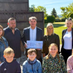 Eine Gruppe von Erwachsenen und Kindern steht bei sonnigem Wetter vor einem Denkmal im Freien. Ein Mann in der Mitte hält einen symbolischen Spendenscheck in den Händen. Alle lächeln in die Kamera.