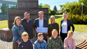 Eine Gruppe von Erwachsenen und Kindern steht bei sonnigem Wetter vor einem Denkmal im Freien. Ein Mann in der Mitte hält einen symbolischen Spendenscheck in den Händen. Alle lächeln in die Kamera.