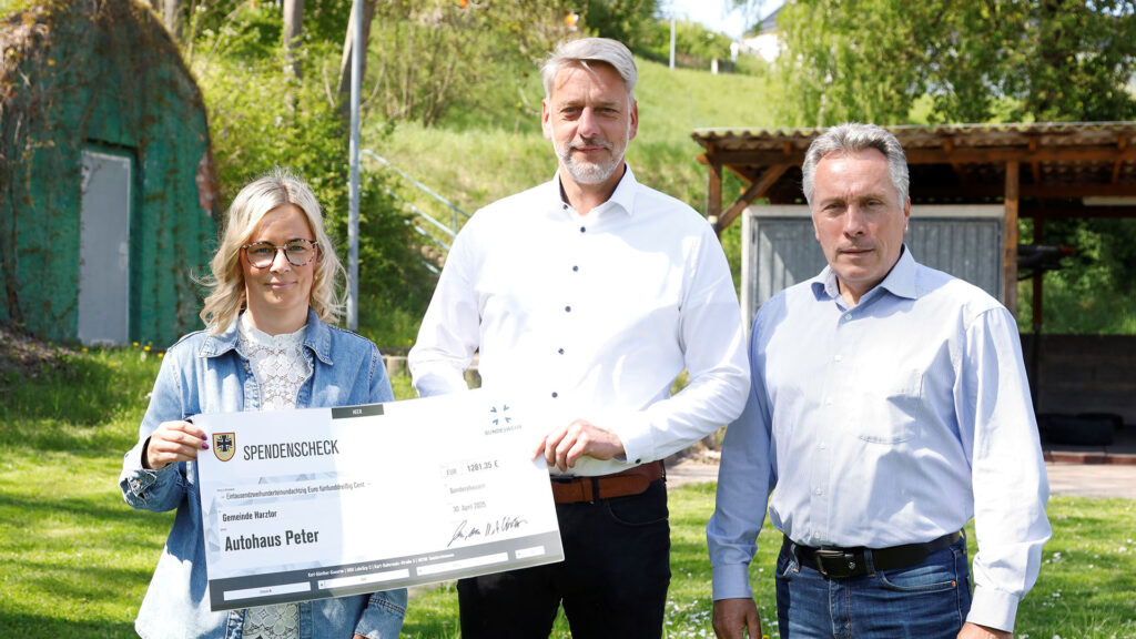 Drei Erwachsene stehen im Freien und halten gemeinsam einen symbolischen Spendenscheck in die Kamera; im Hintergrund sind Bäume, Rasen und ein kleines Gebäude zu sehen.