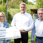 Drei Erwachsene stehen im Freien und halten gemeinsam einen symbolischen Spendenscheck in die Kamera; im Hintergrund sind Bäume, Rasen und ein kleines Gebäude zu sehen.