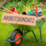 Eine grüne Schubkarre voller Gartenwerkzeuge und Blumentöpfe steht auf einer Wiese, daran befestigt ein Pappschild mit der handgeschriebenen Aufschrift „ARBEITSEINSATZ“ – im Hintergrund ein blühender Garten. KI.