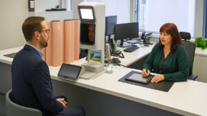Eine Mitarbeiterin im Bürgerbüro nimmt biometrische Passbilder einer Besucherin auf, beide sitzen sich an einem modern ausgestatteten Schalter gegenüber. KI.