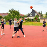 Eine Gruppe Jugendlicher spielt Basketball auf einem roten Freiplatz in einem Wohngebiet, der Ball ist gerade in der Luft. Im Hintergrund sind Häuser und Bäume zu sehen.