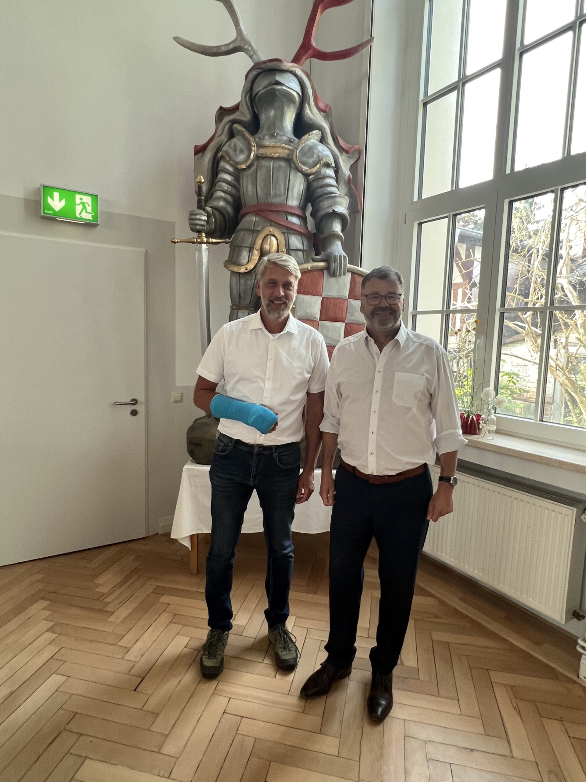 Frank Rostek (links, mit blauem Gipsarm) und Stephan Klante (rechts) stehen nebeneinander in einem hellen Raum mit Holzfußboden. Hinter ihnen eine große Ritterfigur mit Schwert, rechts große Fensterfront mit Blick ins Grüne.