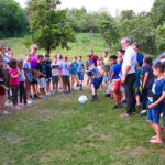 Eine große Gruppe von Kindern und einigen Erwachsenen steht im Halbkreis auf einer Wiese; ein Junge tritt gerade gegen einen Ball. Im Hintergrund sind Bäume, Zelte und eine grüne Hügellandschaft zu sehen.