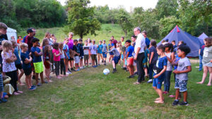 Eine große Gruppe von Kindern und einigen Erwachsenen steht im Halbkreis auf einer Wiese; ein Junge tritt gerade gegen einen Ball. Im Hintergrund sind Bäume, Zelte und eine grüne Hügellandschaft zu sehen.