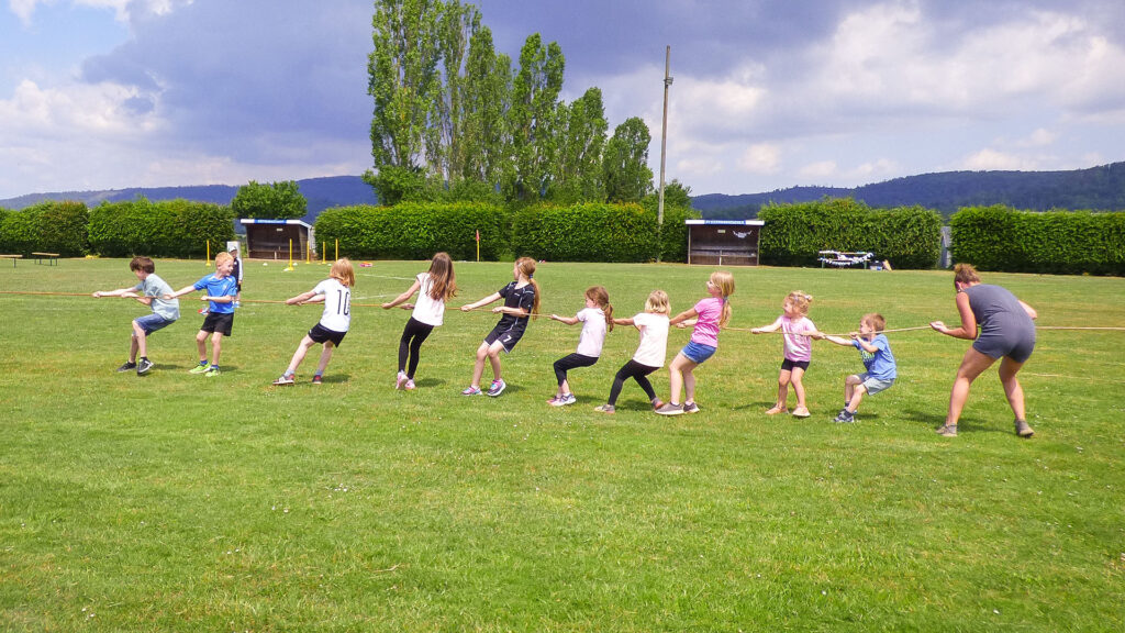 Kinder und eine Erwachsene spielen Tauziehen auf einer grünen Wiese bei einem Sportfest oder Sommerfest, im Hintergrund Bäume und Berge.