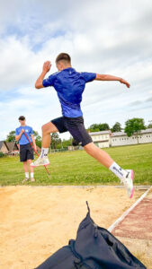 Ein Junge im blauen Sportshirt springt beim Weitsprung über die Sandgrube, während ein anderer Jugendlicher im Hintergrund mit einem Maßstab zur Weitenmessung bereitsteht. Im Hintergrund sind ein Sportplatz, Gebäude und Bäume zu sehen.
