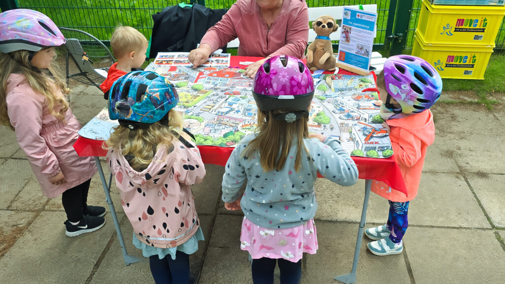 Helmtragende Kinder studieren ein Wimmelbild zum Straßenverkehr an einem Infostand.