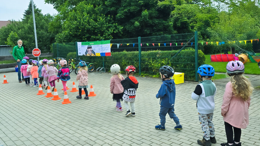 Kinderreihe mit Helmen übt Slalomlauf vor Stopp-Schild und Pylonen.