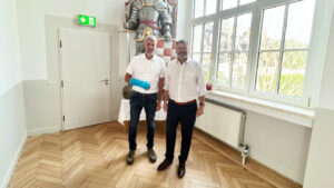 Frank Rostek (links, mit blauem Gipsarm) und Stephan Klante (rechts) stehen nebeneinander in einem hellen Raum mit Holzfußboden. Hinter ihnen eine große Ritterfigur mit Schwert, rechts große Fensterfront mit Blick ins Grüne.