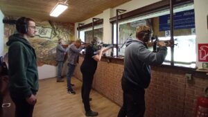 Zwei Schützen in grünen Hoodies stehen nebeneinander in einer Indoor-Schießanlage und zielen mit Kleinkalibergewehren stehend aufgelegt auf fünf runde Klappscheiben; im Hintergrund beobachten mehrere Personen das Geschehen.
