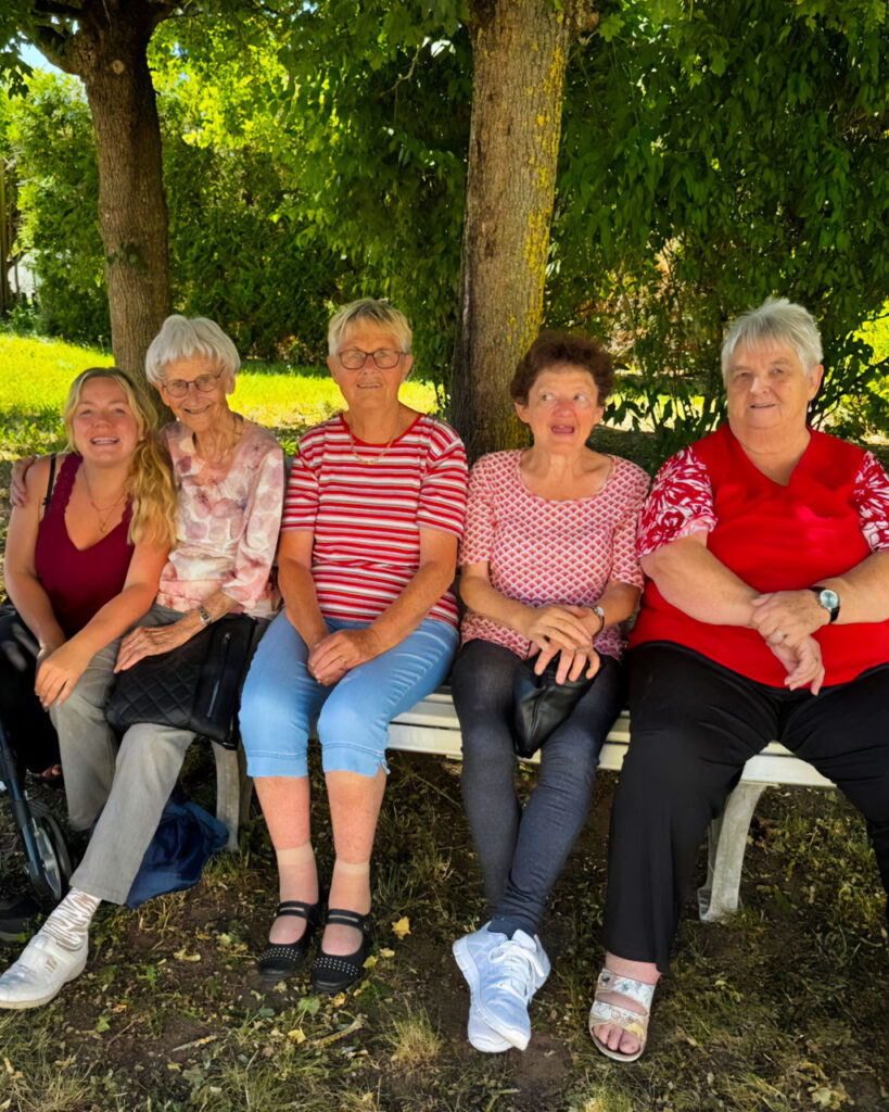 Fünf Frauen unterschiedlicher Generationen sitzen dicht nebeneinander auf einer weißen Bank im Schatten eines großen Baumes, schauen lächelnd in die Kamera; im Hintergrund üppiges Grün.