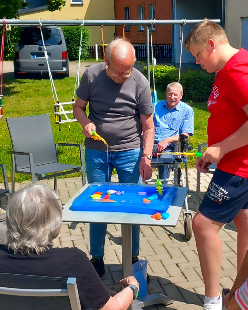 In einem sonnigen Hof spielen mehrere Generationen ein Angelspiel: Ein älterer Mann und ein Jugendlicher halten Spielruten über einen blauen Wasserbehälter mit bunten Plastikfischen auf einem Tisch, während eine Seniorin im Rollstuhl und eine weitere sitzende Person zusehen.