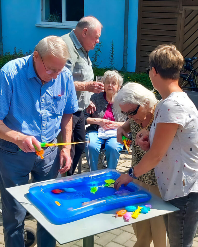 Drei Personen – ein älterer Mann, eine ältere Frau mit Sonnenbrille und eine jüngere Betreuerin – stehen im Freien um einen kleinen, wassergefüllten, blauen Spielpool auf einem Tisch und angeln mit bunten Spielruten nach Plastikfischen.