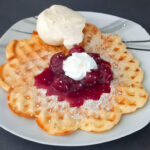 Eine goldbraun gebackene, herzförmige Waffel liegt auf einem weißen Teller; darauf in der Mitte ein Klecks leuchtend roter Kirschsoße mit geschlagener Sahne, rundherum Puderzucker. Oben auf der Waffel ruht eine Kugel Vanilleeis.