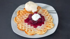 Eine goldbraun gebackene, herzförmige Waffel liegt auf einem weißen Teller; darauf in der Mitte ein Klecks leuchtend roter Kirschsoße mit geschlagener Sahne, rundherum Puderzucker. Oben auf der Waffel ruht eine Kugel Vanilleeis.