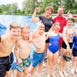 Eine fröhliche Gruppe von neun Kindern in Badebekleidung steht am Beckenrand eines Freibads, winkt lachend in die Kamera und trägt teilweise Schwimmbrillen in der Hand. Hinter ihnen posieren zwei betreuende Erwachsene im T-Shirt – alle vor der Kulisse eines großen Außenpools, grüner Bäume und eines Rutschenauslaufs.