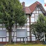 Außenansicht des zukünftigen Gästehauses „Pension Zum Handwagen“ in Niedersachswerfen: weißes Fachwerkhaus mit rotem Ziegeldach, flankiert von zwei Laubbäumen; Logo der Pension oben rechts eingeblendet
