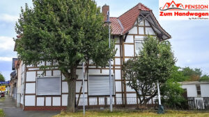 Außenansicht des zukünftigen Gästehauses „Pension Zum Handwagen“ in Niedersachswerfen: weißes Fachwerkhaus mit rotem Ziegeldach, flankiert von zwei Laubbäumen; Logo der Pension oben rechts eingeblendet