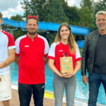 Gruppenfoto am Beckenrand des Waldbades: Shayenne Kara mit Geschenk in der Mitte, links flankiert von Schwimmmeister Roland Gerlach und seinem Kollegen Steven Klante sowie rechts Bürgermeister Stephan Klante – im Hintergrund die blaue Rutsche des Freibades Niedersachswerfen.