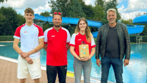 Gruppenfoto am Beckenrand des Waldbades: Shayenne Kara mit Geschenk in der Mitte, links flankiert von Schwimmmeister Roland Gerlach und seinem Kollegen Steven Klante sowie rechts Bürgermeister Stephan Klante – im Hintergrund die blaue Rutsche des Freibades Niedersachswerfen.