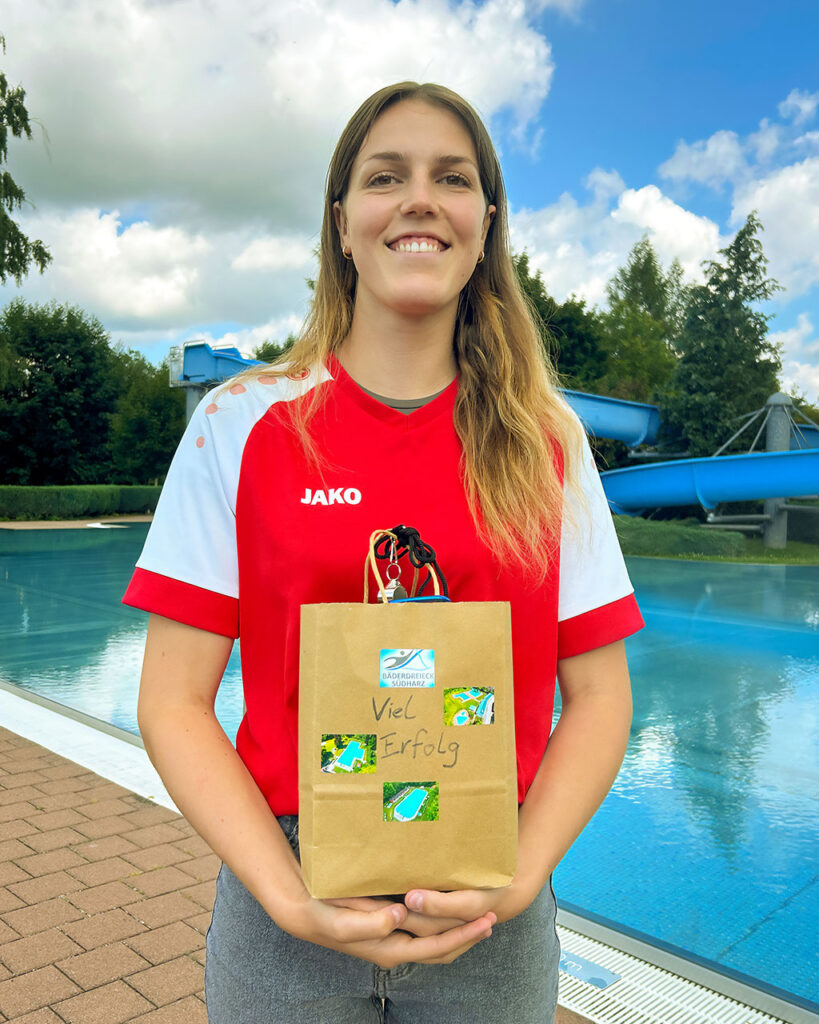 Shayenne Kara lächelt in die Kamera und hält eine Geschenktüte mit der Aufschrift ‚Viel Erfolg‘ vor dem Becken des Freibades Niedersachswerfen – im Hintergrund die markante blaue Rutsche.