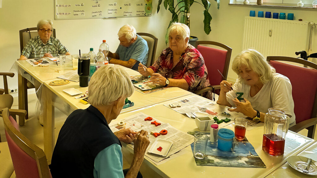 Mehrere Seniorinnen und Senioren sitzen gemeinsam an einem Basteltisch und bemalen bunte Buchstaben aus Ton – konzentrierte Atmosphäre im Gruppenraum.