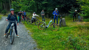 Kurze Pause am Waldrand: lachende Radlerin vorn, dahinter Teilnehmende mit E-Bikes und Mountainbikes auf einem Forstweg.